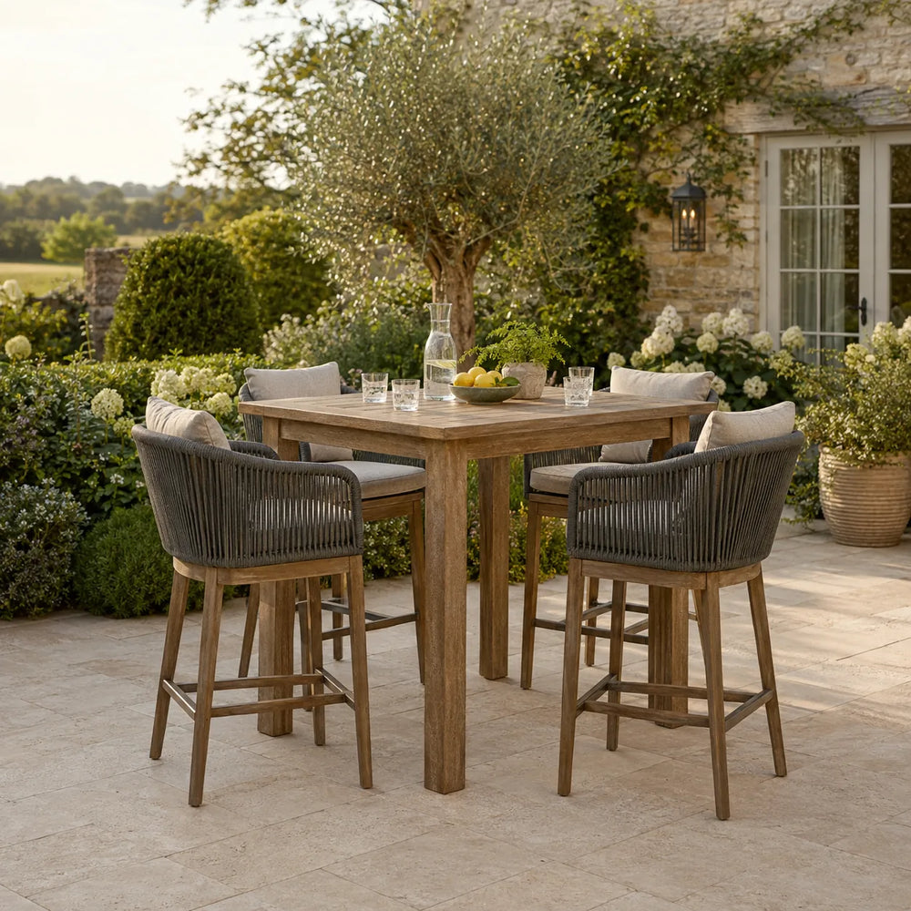 Luxury wooden outdoor bar table with four woven bar stools and cream cushions on a landscaped stone garden terrace – Verdant Lounge UK