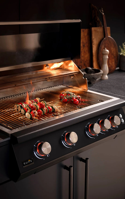 Close-up of the F4 outdoor gas grill with stainless steel cooking grates, five illuminated control knobs, vegetables grilling over open flames and matte black cabinetry.
