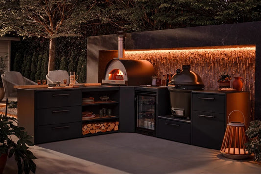 Outdoor kitchen setup at dusk with dark charcoal L-shaped cabinets, a wood-fired pizza oven with visible flames, a black grill, a small glass-door refrigerator, wood storage, and warm ambient lighting against a textured wall.