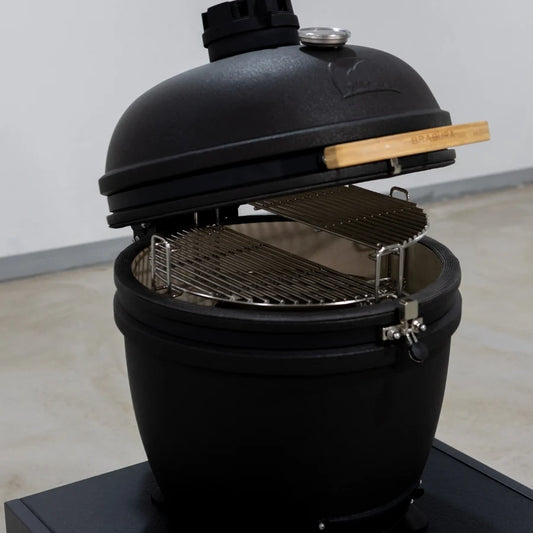 A black ceramic kamado grill with its lid partially open, showing stainless steel racks inside, photographed in a clean indoor setting.