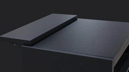 A close-up of a dark gray textured surface with a rectangular black lid or cover resting on top, set against a dark studio background/Verdant Lounge.
